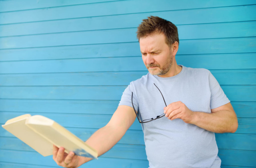 A farsighted man holding his book in distant while holding his glasses on the other hand