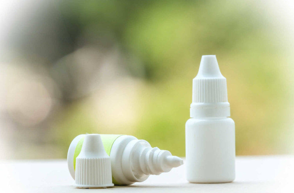Two bottles of eye drops sitting on a counter, one standing up and one on it's side with the cap off in front of a green background
