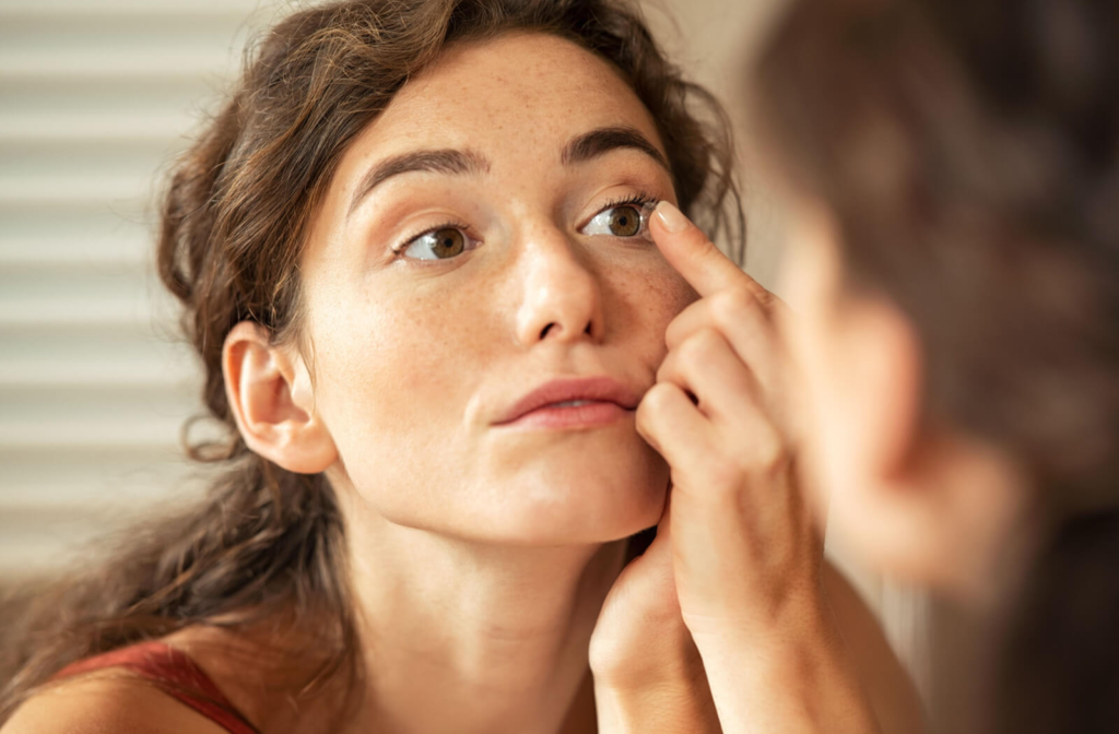 A young woman standing in front of a mirror and putting a contact lens on her left eye.