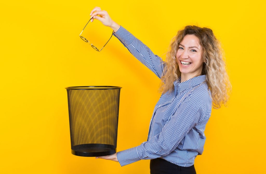 A smiling woman throws her glasses in the garbage.