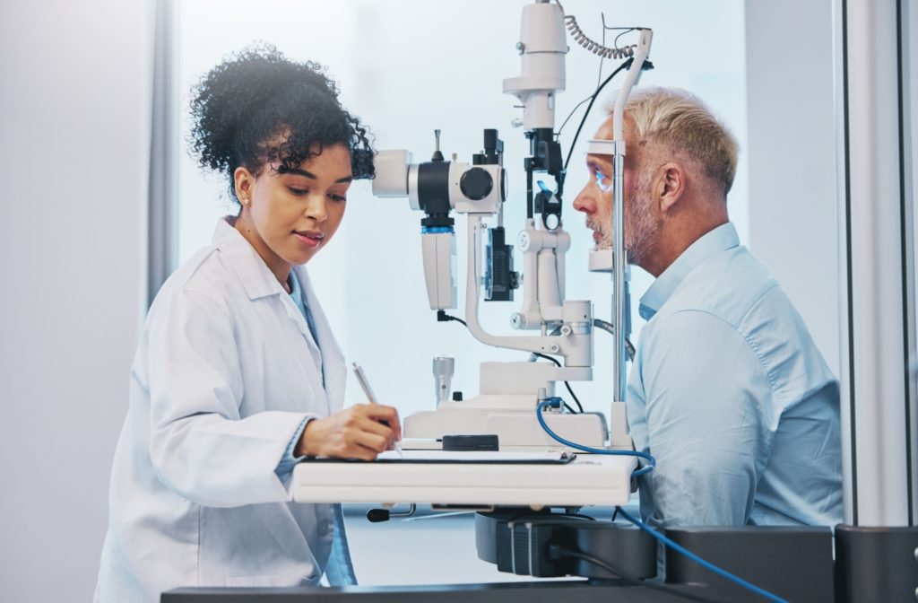 An eye doctor writes down the results of an eye examination as the patient sits at a slit lamp.