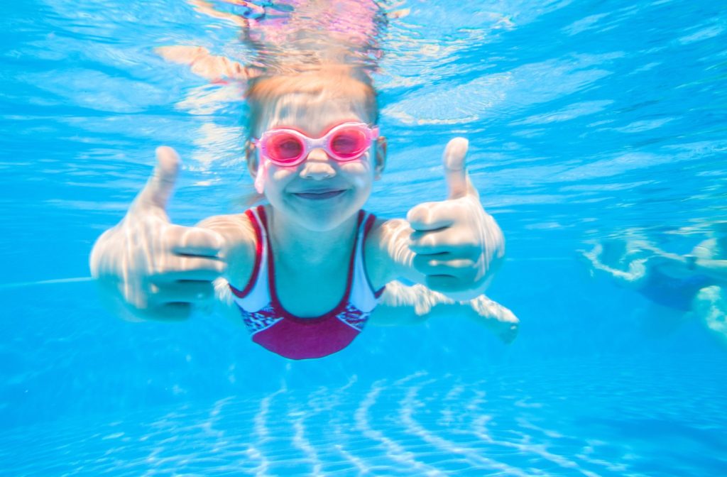 A young girl swimming in a pool and wearing goggles gives two thumbs up.