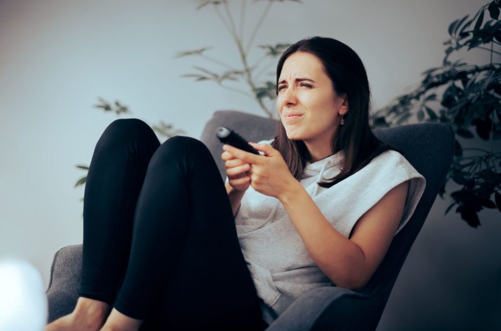 A woman sitting on a couch squinting their eyes from myopia and holding a TV remote in their hands.