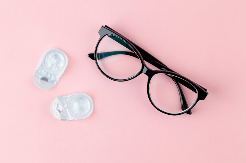 A pair of eyeglasses and contact lenses on a pink background.
