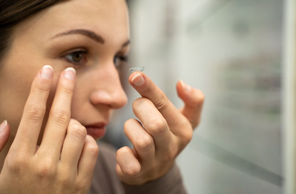 A close-up of a patient using their right hand to gently pull down the lower eyelid of their right eye while preparing to insert a contact lens with their left hand.