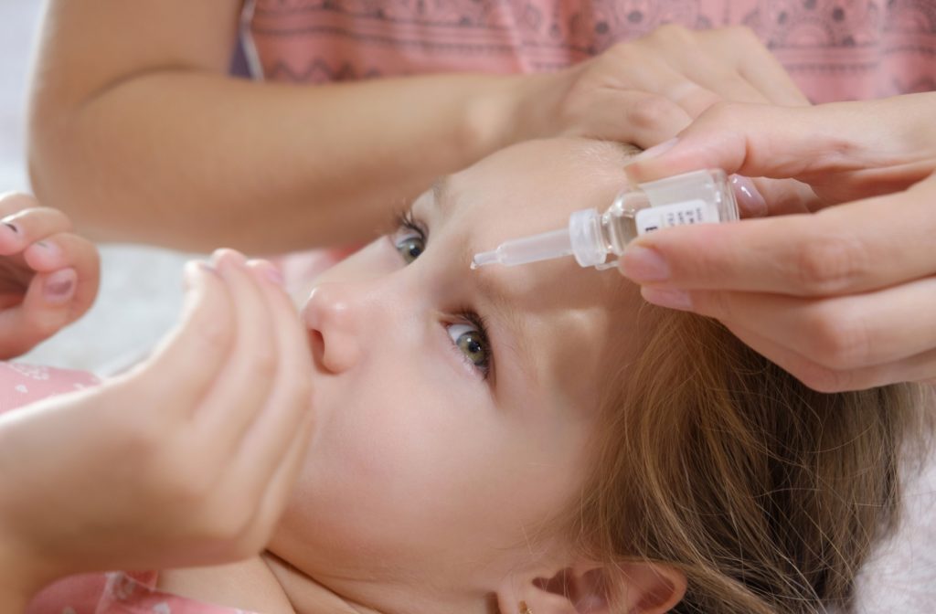 A child lies on their back, looking up in concern as their parent holds an eye dropper over them to administer medicine