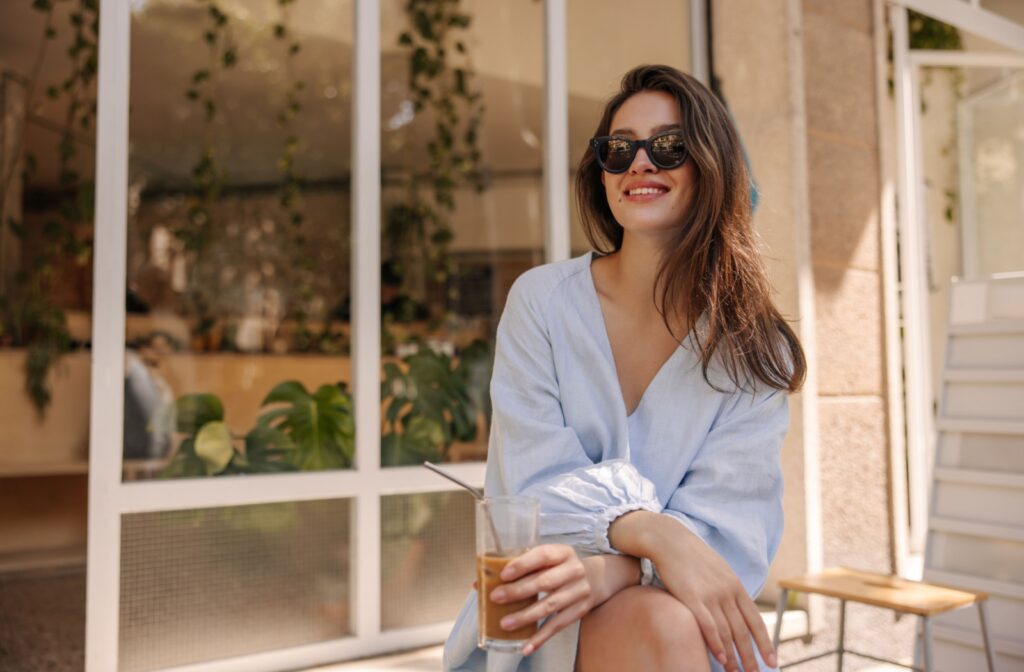 A woman sitting outside of a cafe, drinking an iced coffee, wearing sunglasses to protect her eyes from the sun's UV rays.