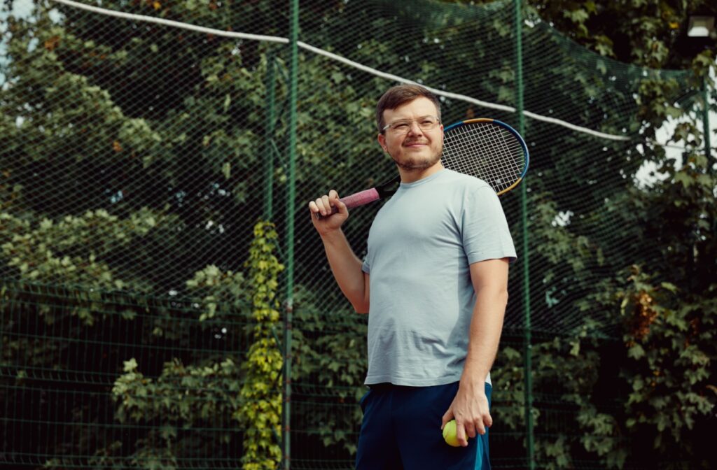 A person outside on a tennis court, holding a tennis racket over their shoulder while wearing sport specific eyeglasses.