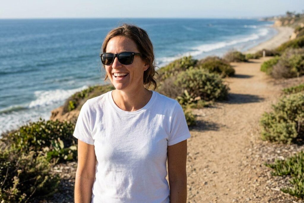Smiling adult wearing sunglasses and a white t-shirt standing on a coastal cliff trail with the ocean and shoreline in the background.